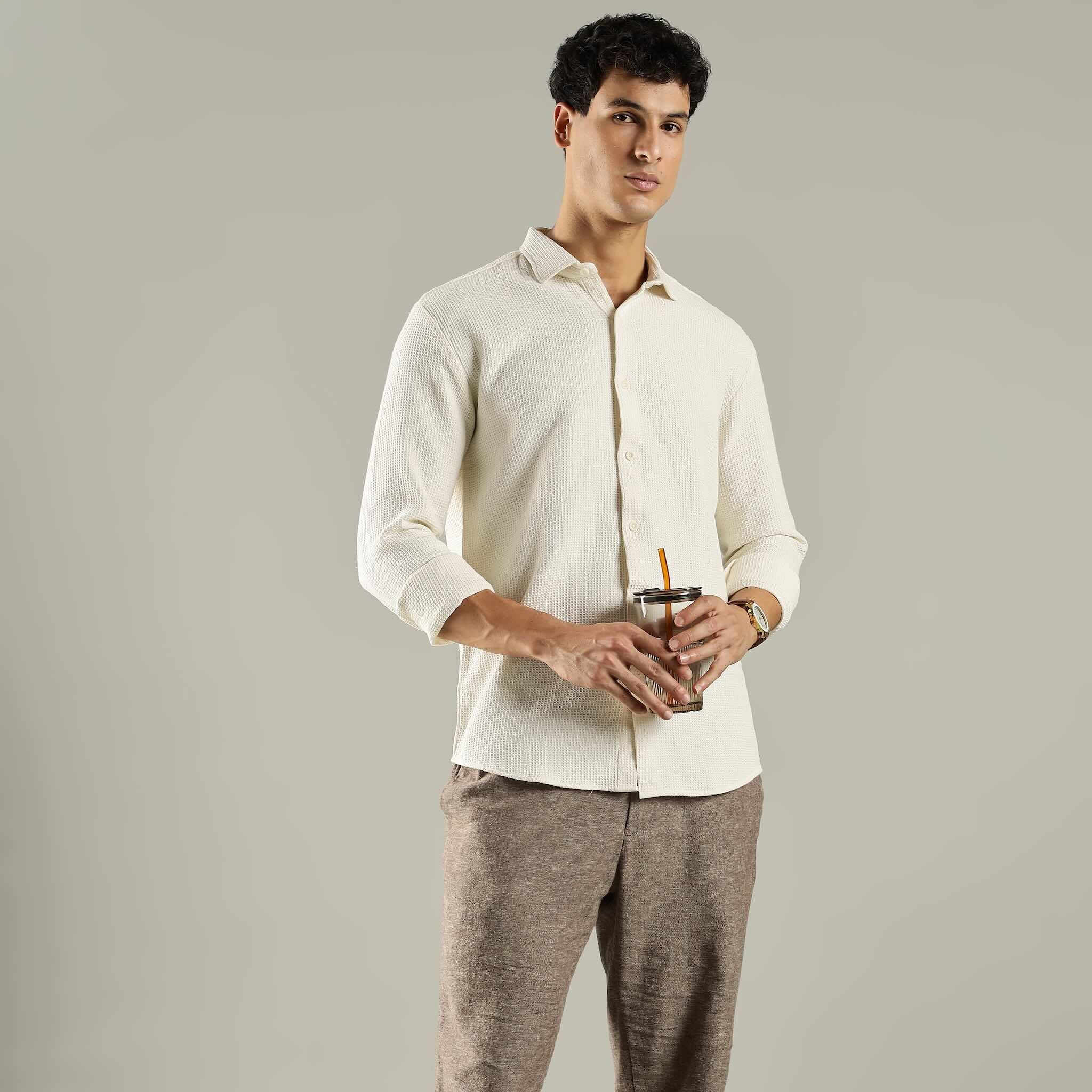 Man wearing a white shirt and brown pants holding a drink against a gray background