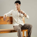 Man sitting on a wooden bench holding a glass of milk, wearing a white shirt and brown pants.