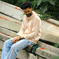 Man wearing sandstone shirt and sitting on concrete steps wearing a beige shirt and blue jeans, with a green bag beside him.