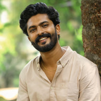Man wearing sandstone shirt with a beard and mustache smiling outdoors, leaning against a tree