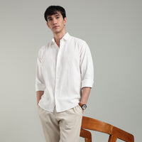 Man wearing a white hemp shirt and beige pants leaning on a chair against a gray background
