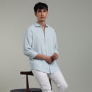 Man wearing a light blue shirt and white pants leaning  on a chair against a gray background