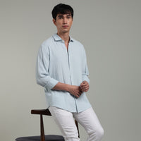 Man wearing a light blue shirt and white pants leaning  on a chair against a gray background