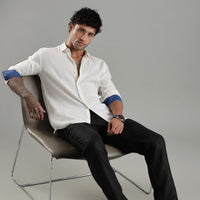 Man sitting on a chair wearing a white shirt with rolled-up sleeves and black pants against a gray background