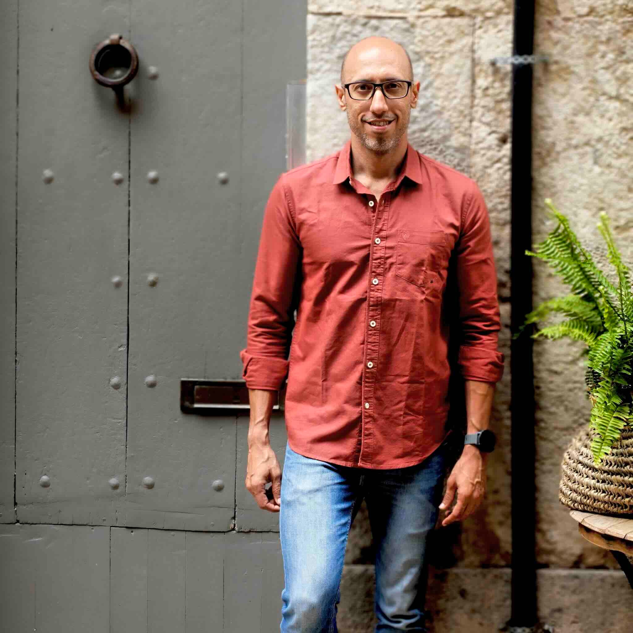 Man wearing a red shirt and blue jeans standing in front of a textured wall and metal door.