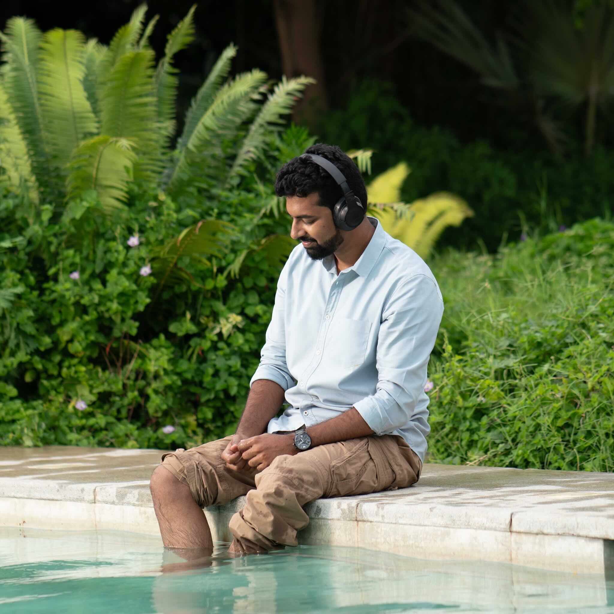 Man wearing a light blue shirt and is sitting by a pool with headphones on, surrounded by greenery