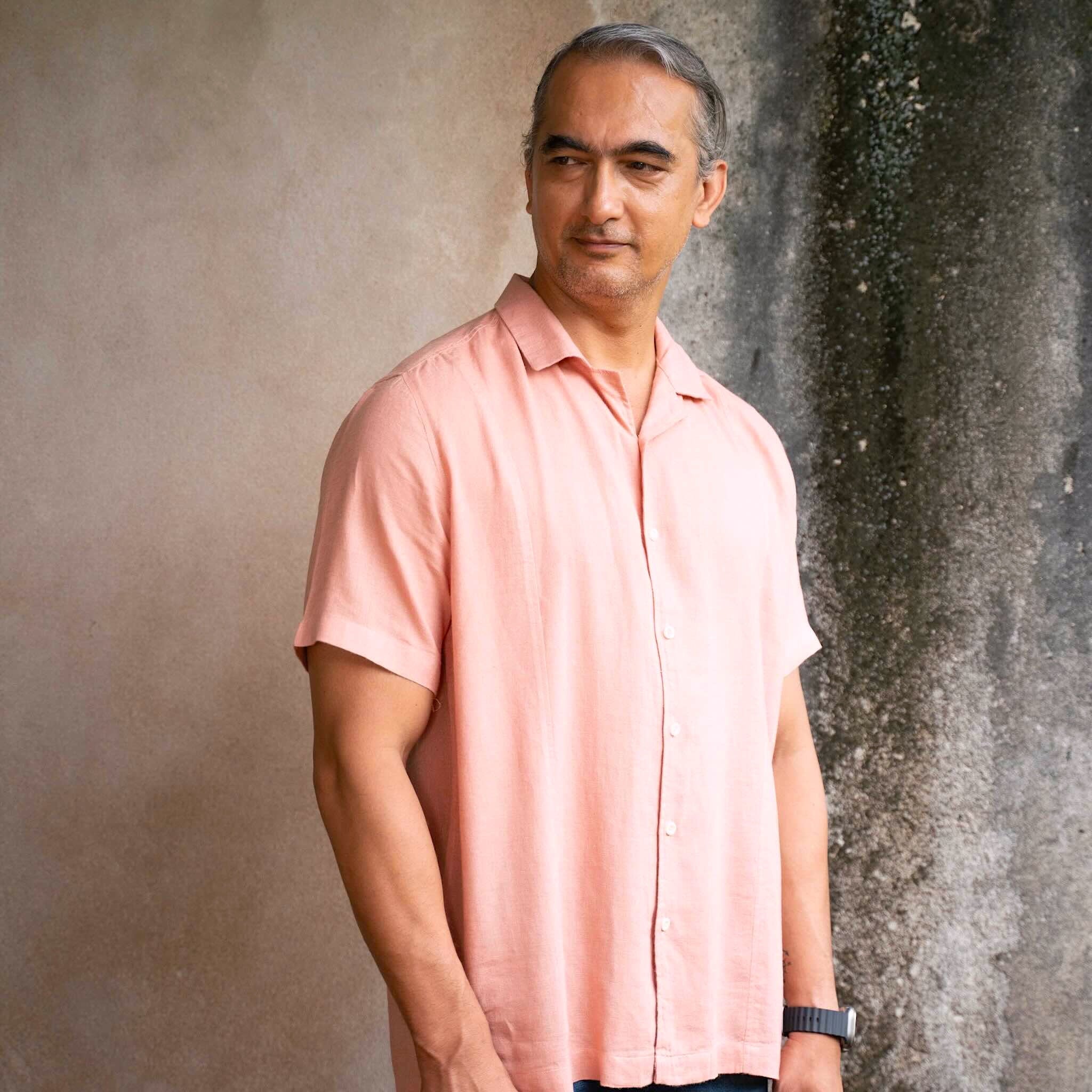Man wearing a light pink shirt standing against a textured wall.